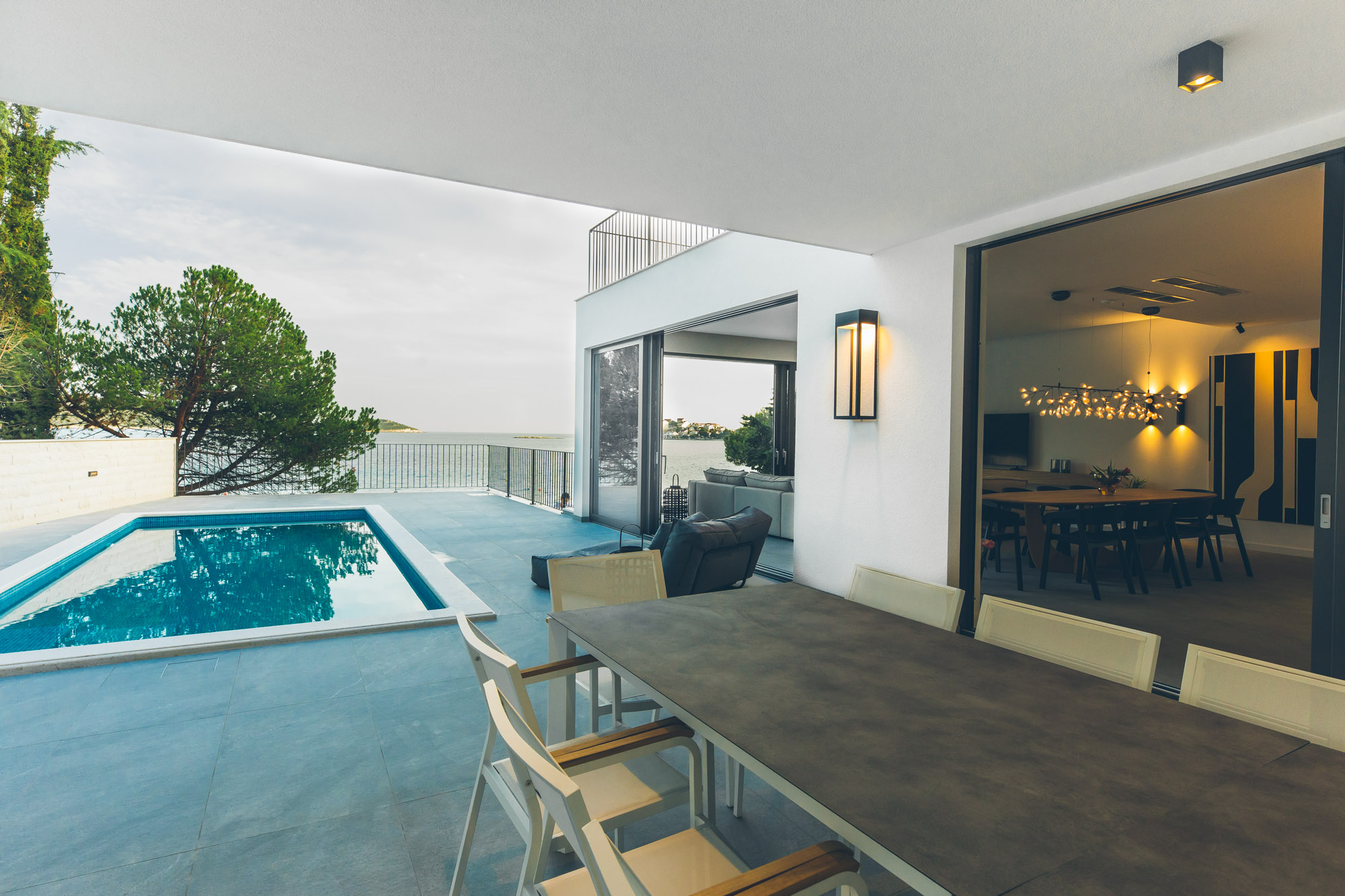 Villa exterior at dusk — pool terrace, outdoor dining, living room glowing inside